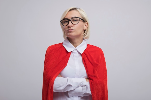 Confident middle-aged blonde superhero woman in red cape wearing glasses standing with closed posture  isolated on white wall with copy space