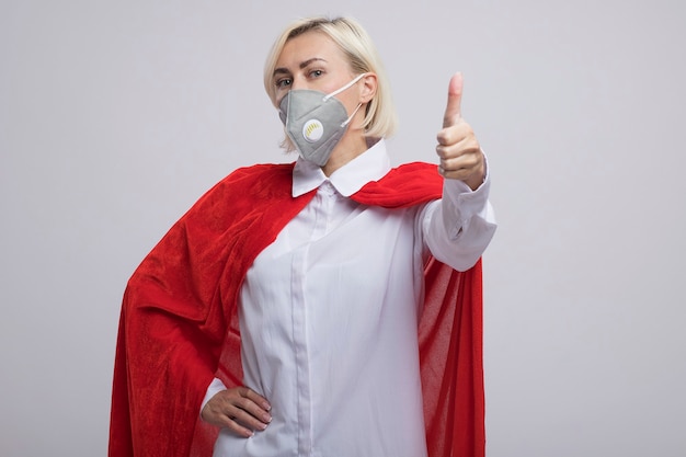 Donna supereroe bionda di mezza età sicura in mantello rosso che indossa una maschera protettiva tenendo la mano sulla vita guardando la parte anteriore che mostra pollice in su isolato sul muro bianco con spazio di copia