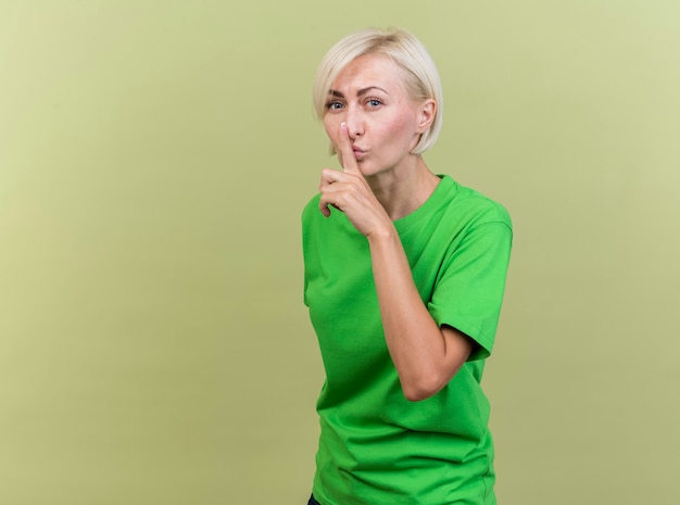 Confident middle-aged blonde woman standing in profile view looking at front doing keep silence gesture isolated on olive green wall