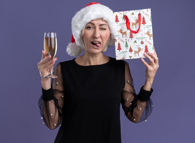 Confident middle-aged blonde woman wearing christmas hat looking at camera holding glass of champagne and touching head with christmas gift bag winking and showing tongue isolated on purple background