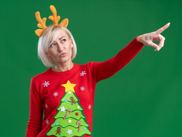 Confident middle-aged blonde woman wearing christmas reindeer antlers headband and christmas sweater looking and pointing at side with pursed lips isolated on green background