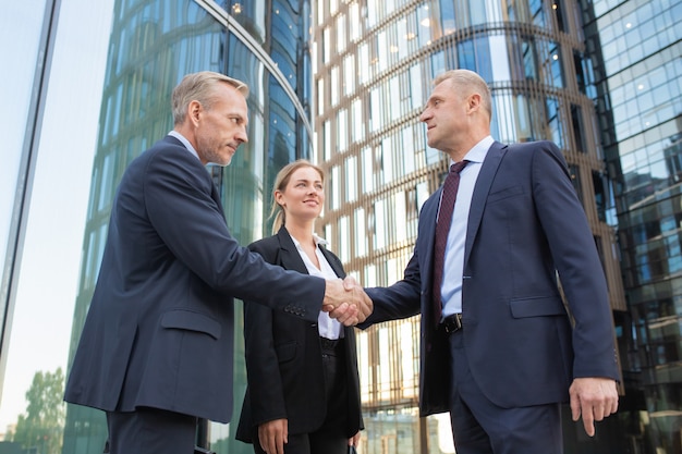 Confident middle-aged businessmen greeting each other and handshaking. Successful content young businesswoman standing near them and smiling. Building on background. Teamwork and partnership concept