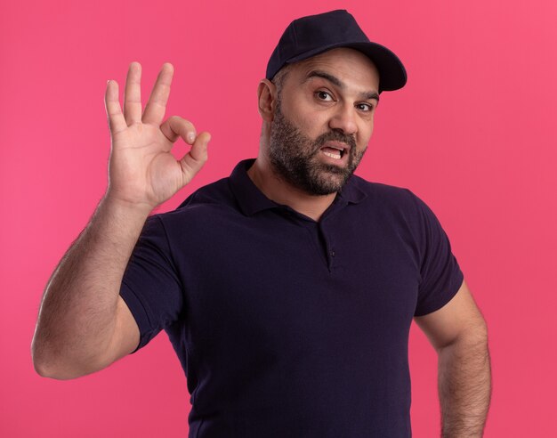 Confident middle-aged delivery man in uniform and cap showing okay gesture isolated on pink wall