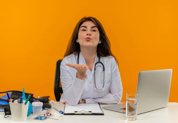 Confident middle-aged female doctor wearing medical robe and stethoscope sitting at desk with medical tools clipboard and laptop looking and sending blow kiss isolated