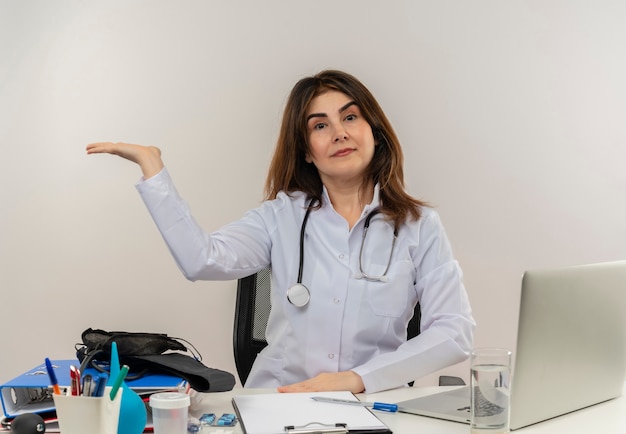 Уверенная женщина-врач средних лет в медицинском халате и стетоскопе, сидя за столом с медицинскими инструментами, буфером обмена и ноутбуком, показывая пустую изолированную руку