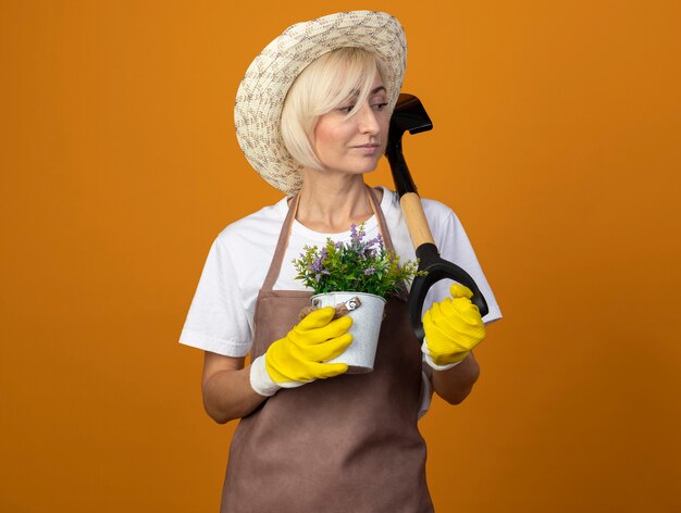 Confident middle-aged gardener woman in gardener uniform wearing hat and gardening gloves holding flowerpot and spade looking at side 