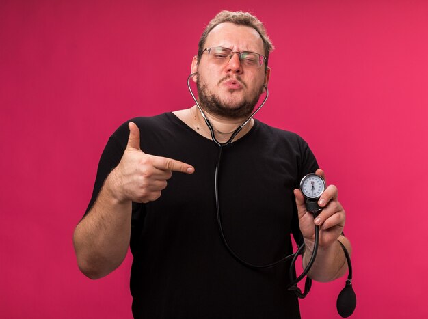 Confident middle-aged ill male measuring his own pressure with sphygmomanometer points at camera 