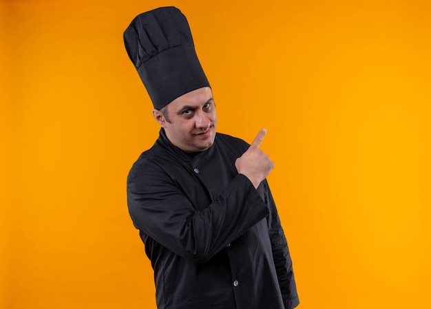 Free photo confident middle-aged male cook in chef uniform points to side on yellow wall with copy space