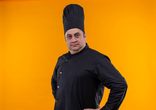 Confident middle-aged male cook in chef uniform put hands on hip on yellow wall with copy space