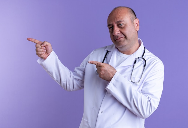confident middle-aged male doctor wearing medical robe and stethoscope looking at front pointing at side isolated on purple wall