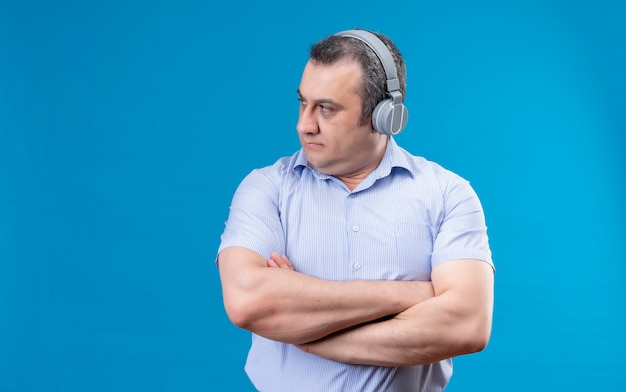 Confident middle-aged man in blue striped shirt wearing headphones with folded arms looking to the right side on a blue space