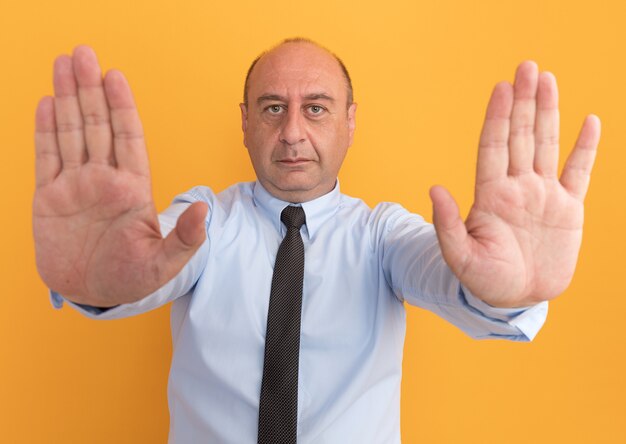 Confident middle-aged man wearing white t-shirt with tie holding out hands at front isolated on orange wall