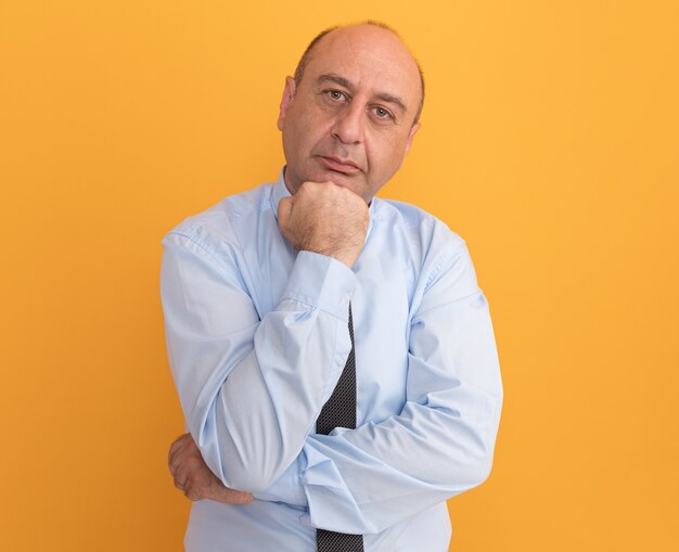 Уверенный в себе мужчина средних лет в белой футболке с галстуком, держащий кулак под подбородком на оранжевой стене