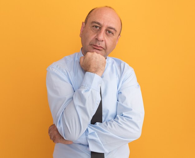 Confident middle-aged man wearing white t-shirt with tie putting fist under chin isolated on orange wall