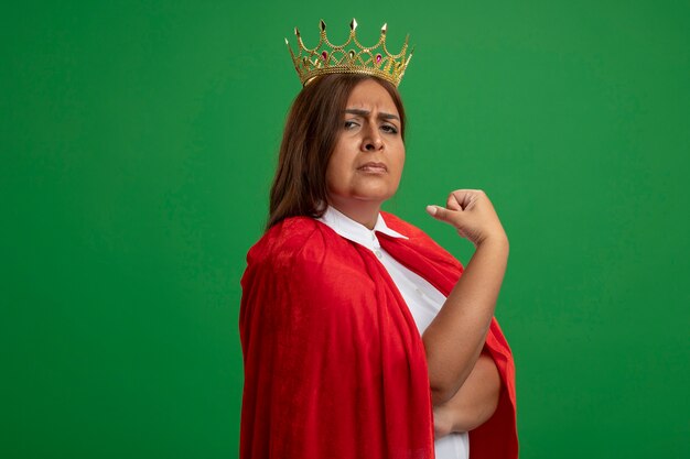 Confident middle-aged superhero female wearing crown points at herself isolated on green