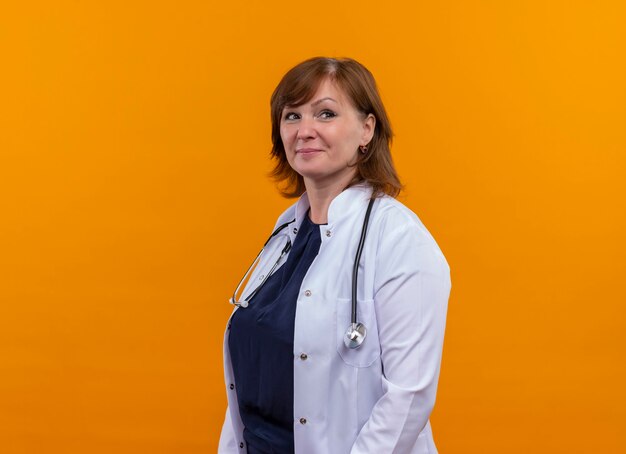 Confident middle-aged woman doctor wearing medical robe and stethoscope standing in profile view on isolated orange wall with copy space