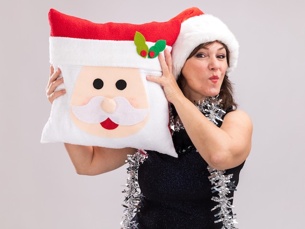 Free photo confident middle-aged woman wearing santa hat and tinsel garland around neck holding santa claus pillow touching head with it looking at camera with pursed lips isolated on white background