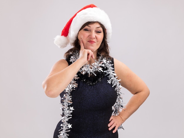 Confident middle-aged woman wearing santa hat and tinsel garland around neck looking at camera keeping hand on waist and on chin isolated on white background