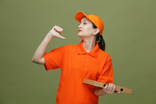 Уверенная в себе женщина-доставщик средних лет в униформе и кепке, держащая пакет с пиццей, смотрит в сторону, указывая на себя, изолированную на оливково-зеленом фоне