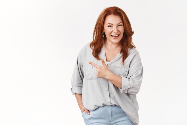 Confident middleaged redhead european woman telling about interesting product recommend checkout promo smiling toothy selfassured pointing left index finger assuring you like it white background