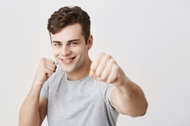 Confident muscular caucasian sportsman with dark hair dressed in gray shirt holding fists in front, going to fight and defend himself. Handsome european guy ready for challenges showing strength