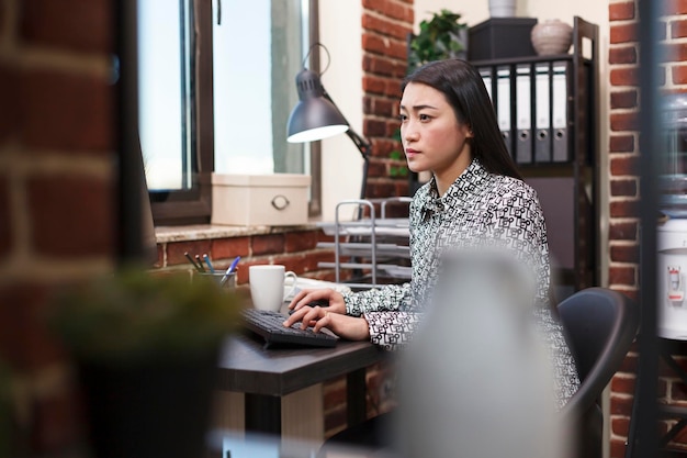 Foto gratuita fiduciosa donna d'ufficio che ricerca idee di marketing di progetto mentre utilizza il computer di lavoro e si siede alla scrivania. donna d'affari dell'agenzia finanziaria che esamina la strategia aziendale mentre si trova nell'area di lavoro.