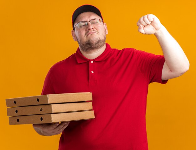 ピザの箱を保持し、コピースペースでオレンジ色の壁に拳を隔離して維持する光学メガネで自信を持って太りすぎの若い配達員