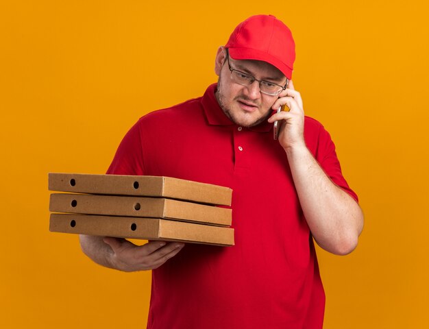 ピザの箱を保持し、コピースペースのあるオレンジ色の壁に隔離された電話で話している光学メガネで自信を持って太りすぎの若い配達員