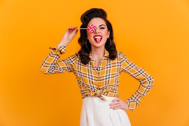 Confident pinup girl posing with pink lollipop. Studio shot of stylish brunette woman in checkered shirt isolated on yellow background.