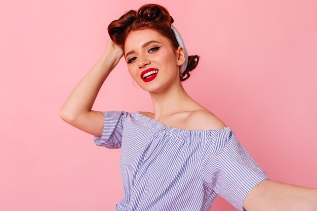 Confident pinup lady making selfie with smile. Studio shot of merry ginger woman isolated on pink space.