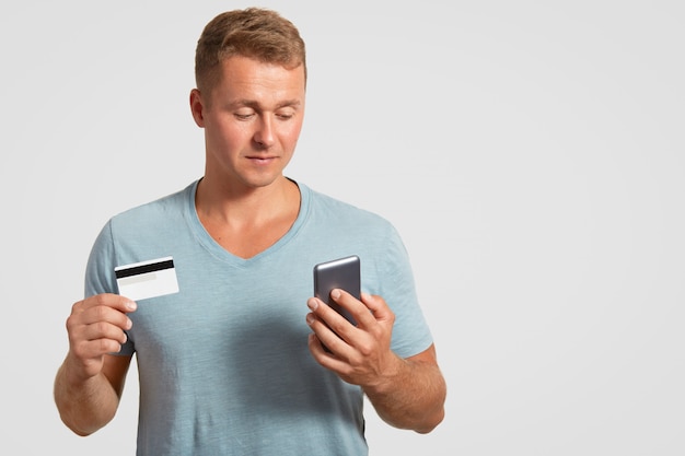 Confident plesant looking male holds modern cellular and plastic card, checks his bank account