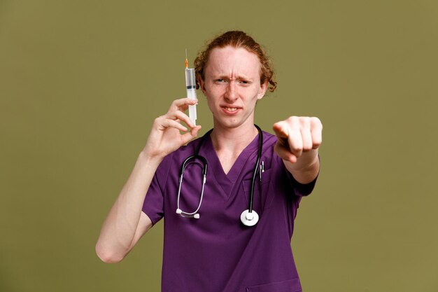 Confident points at camera young male doctor wearing uniform with stethoscope holding syringe isolated on green background