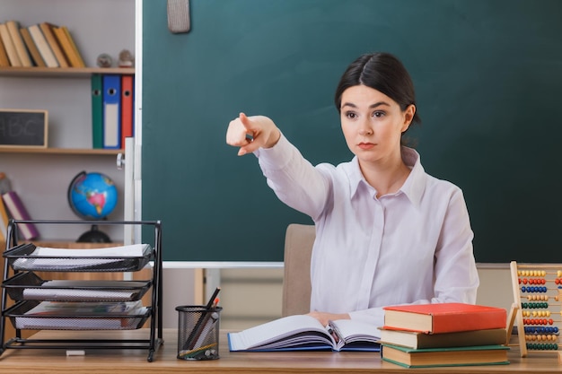 confident points at side young female teacher sitting at desk with school tools in classroom