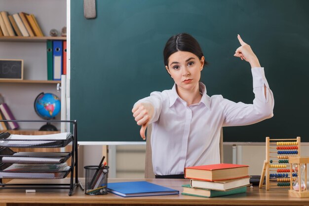 confident points at up showing thumbs down young female teacher sitting at desk with school tools in classroom