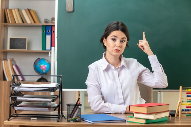 Confident points at up young female teacher sitting at table with school tools in classroom