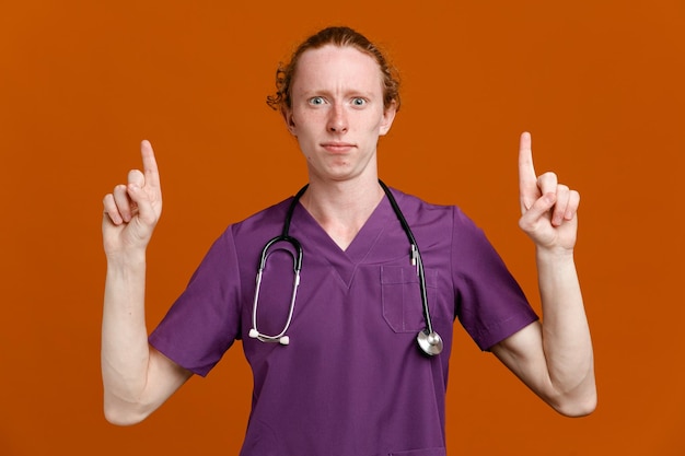 confident points at up young male doctor wearing uniform with stethoscope isolated on orange background