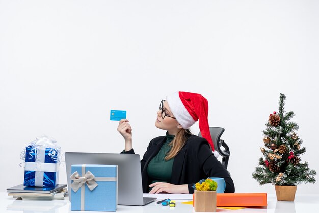 サンタクロースの帽子とテーブルに座って、オフィスで銀行カードを保持している眼鏡を身に着けている自信を持って前向きで忙しい魅力的な女性