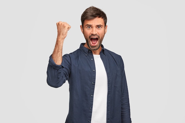 Confident positive male winner keeps hand raised clenched in fist, has widely opened mouth, exclaims with triumph, being emotional, feels success, stands against white wall. Achievement concept