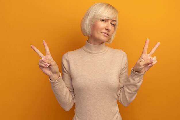 Confident pretty blonde slavic woman gestures victory hand sign with two hands on orange