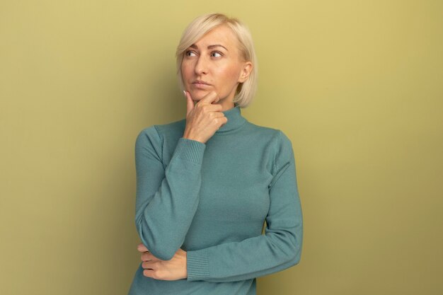 Confident pretty blonde slavic woman holds chin and looks at side on olive green