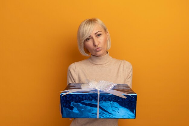 Confident pretty blonde slavic woman holds gift box isolated on orange wall