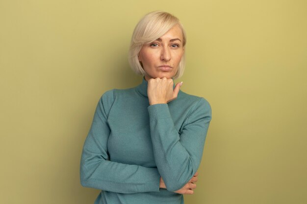 Confident pretty blonde slavic woman puts hand on chin looking at camera on olive green