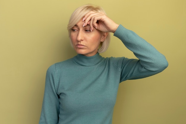 Free photo confident pretty blonde slavic woman puts hand on forehead and looks at side on olive green