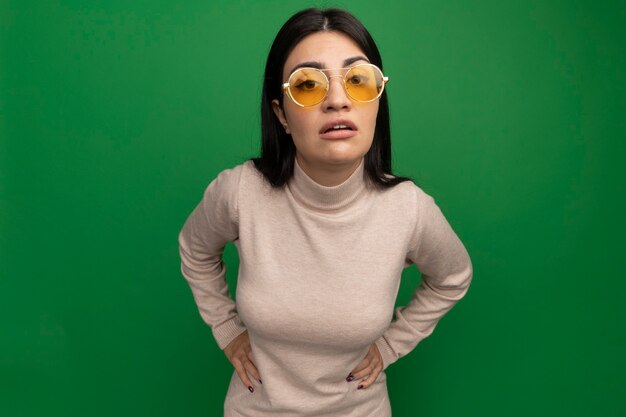 Confident pretty brunette caucasian girl in sun glasses puts hands on waist and looks at camera on green