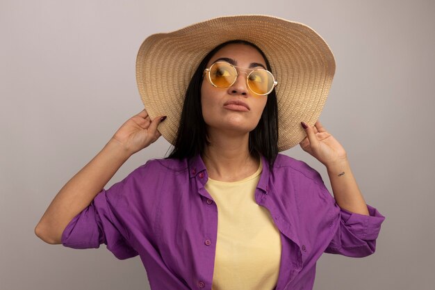 Confident pretty brunette caucasian girl in sun glasses with beach hat looks at side on white