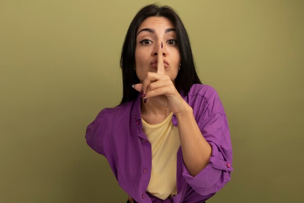 Confident pretty brunette woman doing silence gesture looking at front isolated on olive green wall