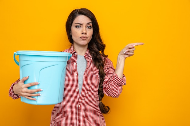 Free photo confident pretty caucasian cleaner woman holding bucket and pointing at side