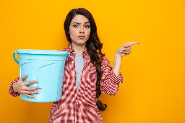 Confident pretty caucasian cleaner woman holding bucket and pointing at side 