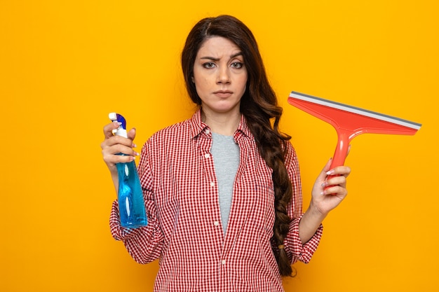 Confident pretty caucasian cleaner woman holding squeegee and spray cleaner and looking at front