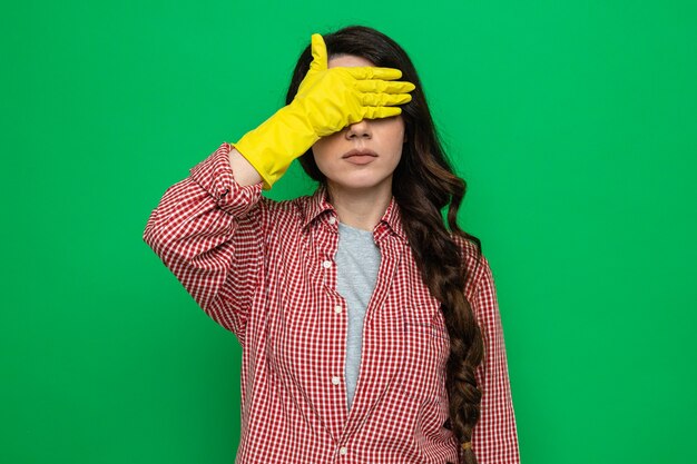 Confident pretty caucasian cleaner woman with rubber gloves covering her eyes with hand